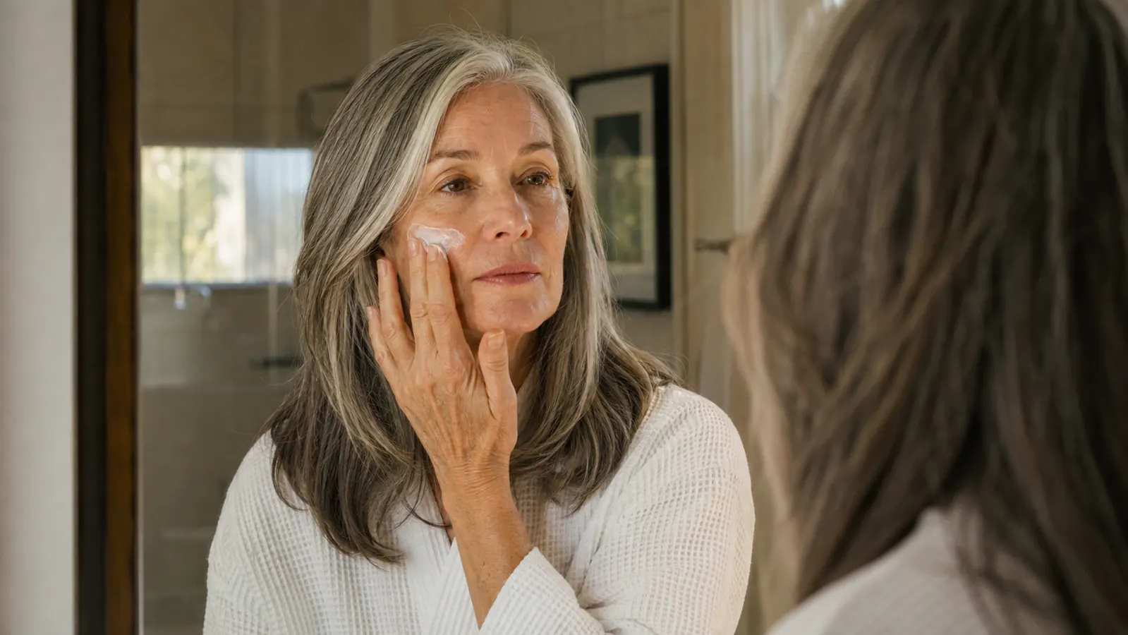 A woman in her early sixties applying moisturizer at a bathroom mirror in soft morning light