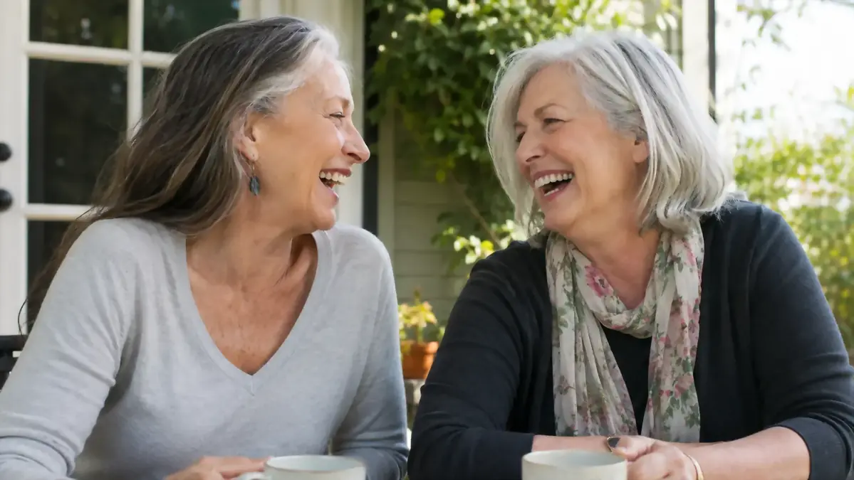 A woman in her sixties wearing a v-neck sweater and smiling with a friend outdoors