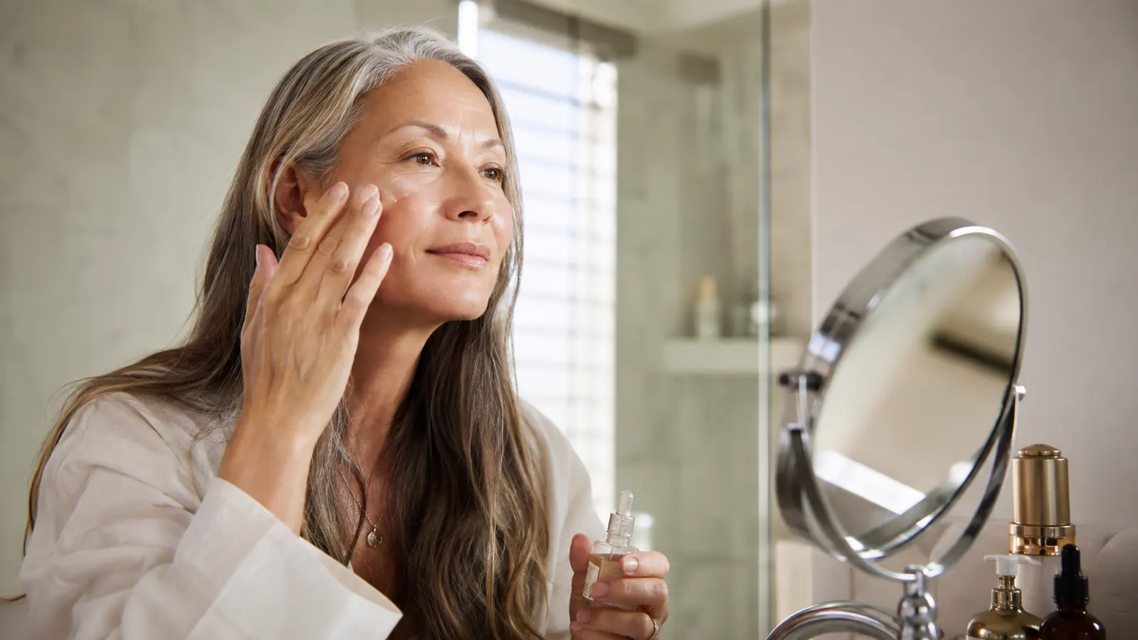 Woman in her late fifties applying skincare serum at a bathroom mirror