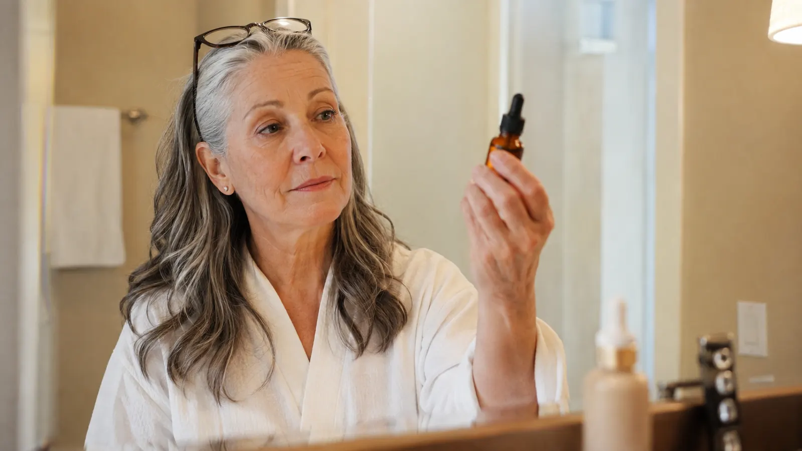 Woman in her sixties standing at a bathroom counter comparing two skincare bottles under warm morning light
