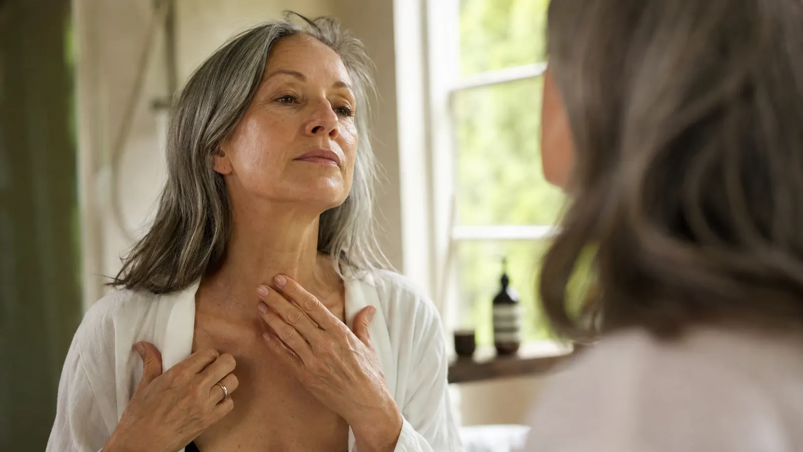 A woman in her sixties looking at her neck and collarbone in a bathroom mirror in soft morning light