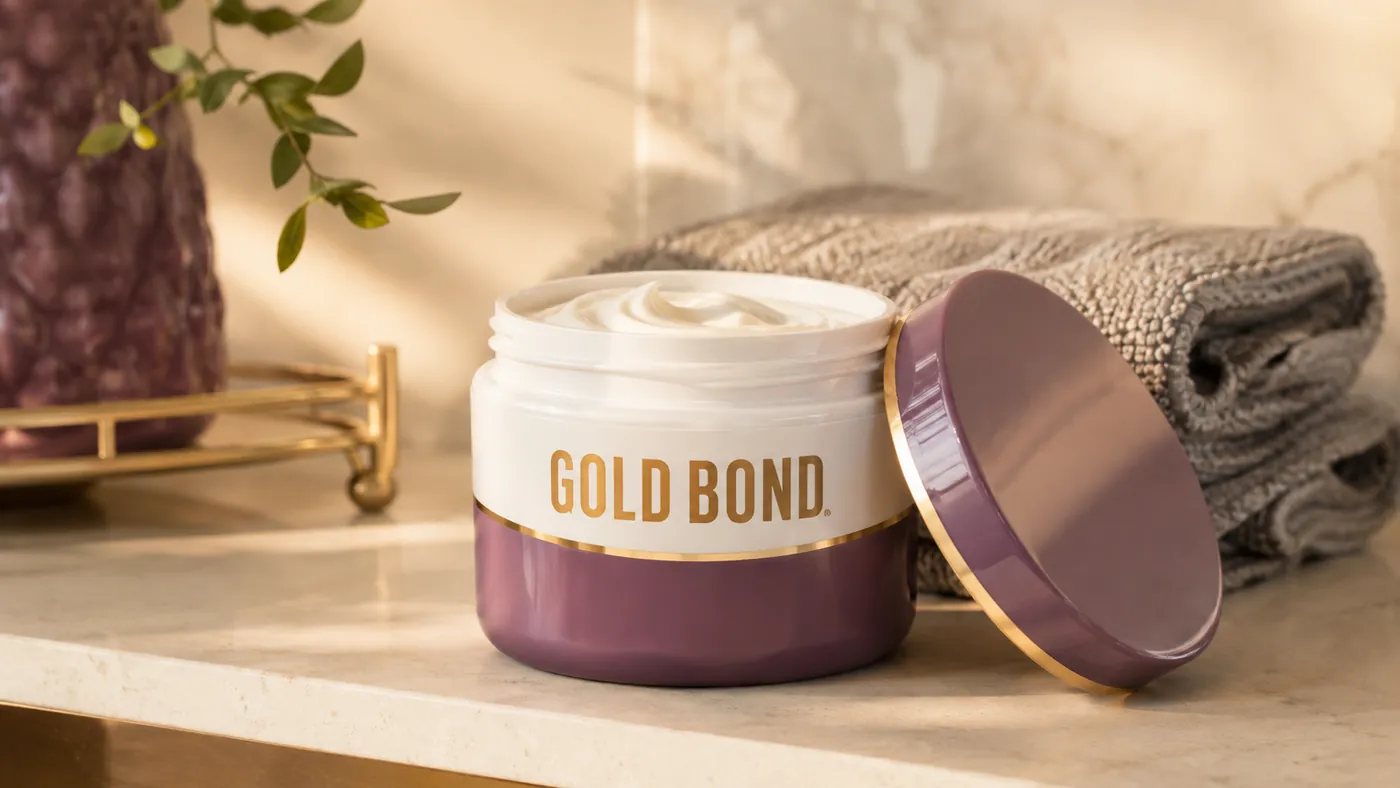 A hand scooping a small amount of Gold Bond Age Renew Neck and Chest Firming Cream from its jar, the Gold Bond wordmark visible on the label