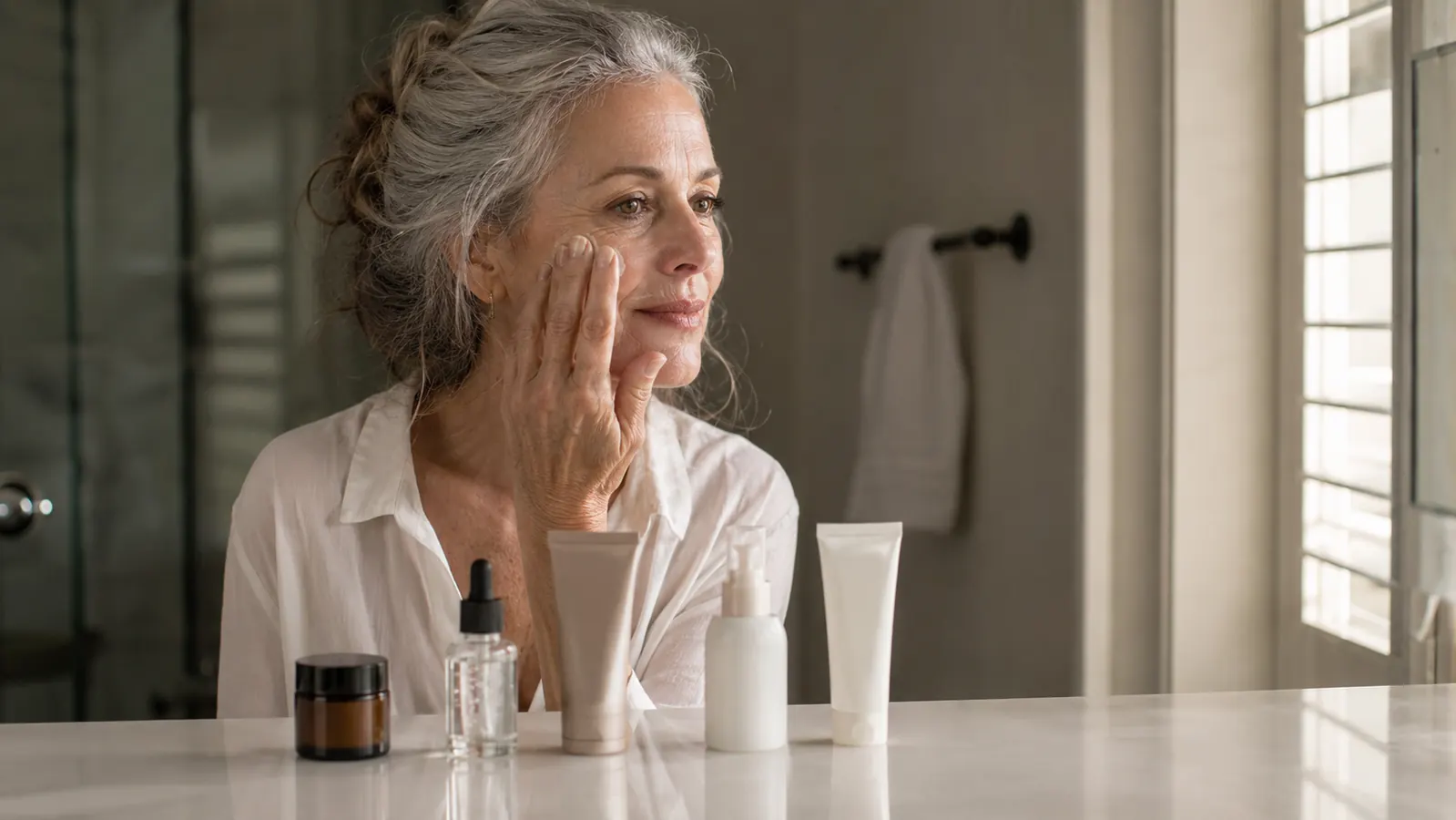 A woman in her fifties standing at a bathroom counter with five simple skincare products lined up, applying cream to her face in soft morning light