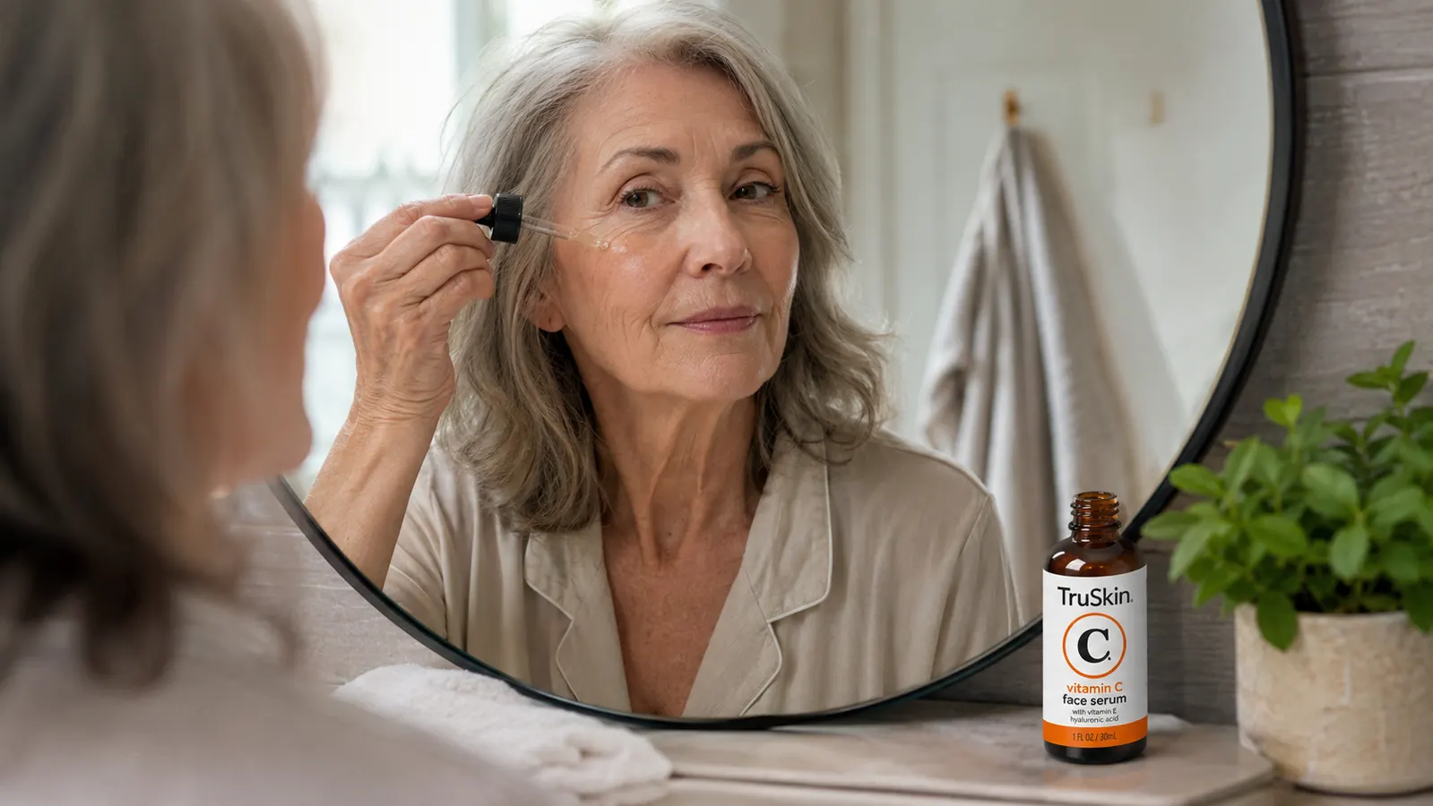 Hand applying vitamin C serum from a dropper to a cheek, with the TruSkin serum bottle visible on the bathroom counter
