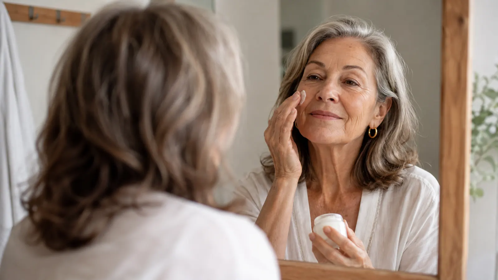 Woman in her sixties applying moisturizer at a bathroom mirror in soft morning light