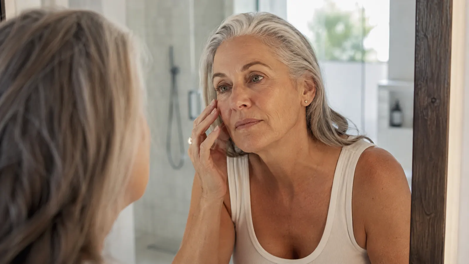 Woman in her early sixties leaning toward a bathroom mirror in soft morning light, checking her cheekbone