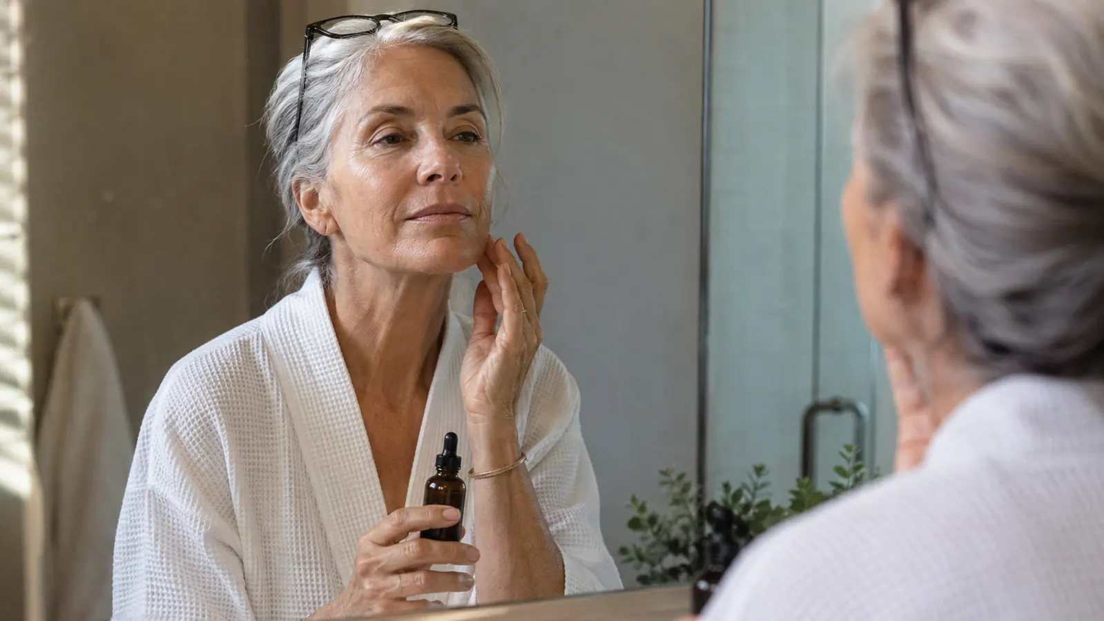 A woman in her sixties standing at a bathroom mirror in soft morning light, holding a small amber serum bottle