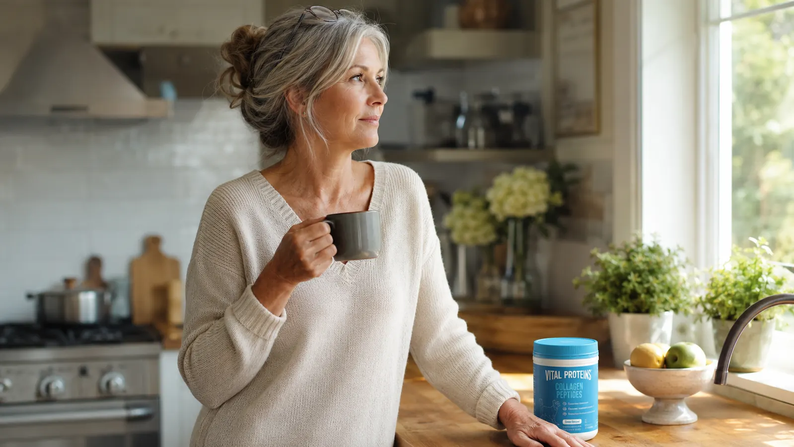 A woman in her sixties standing at a kitchen counter in soft morning light, holding a mug of coffee next to a jar of collagen powder